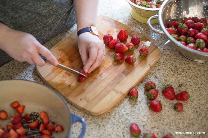 Someone cutting strawberries on a wooden chopping board. 
Chopped strawberries are placed in a large pot and a strainer is full of clean strawberries waiting to be cut.