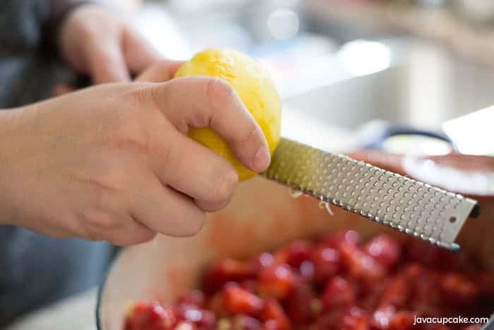 A pair of hand is grating lemon zest on top of the pot of clean cut strawberries.