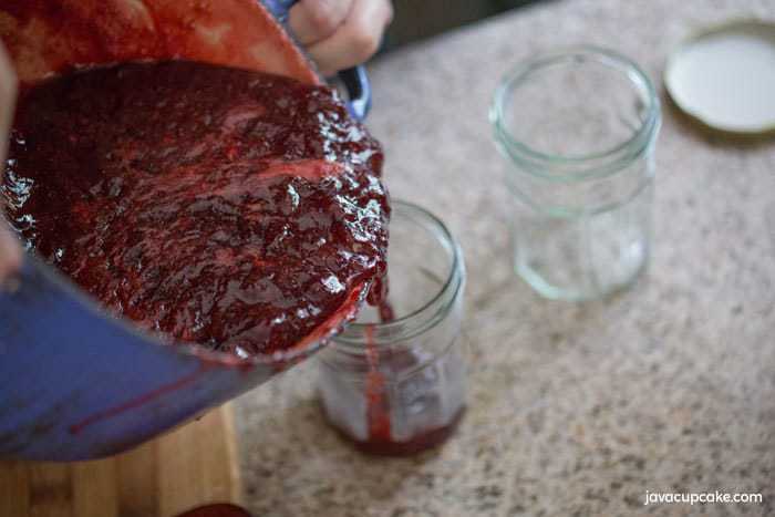 Pouring the completed homemade strawberry preserve from the blue pot into a clean glass jar. Another empty glass jar sits on top of the counter, waiting to be filled.