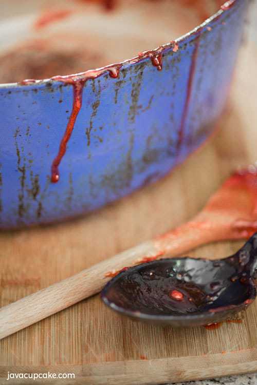Close up shot of pots and ladles used in cooking, civered with sticky red strawberry jam, the jam has dripped down the side of the blue pot and covers the wooden spoon and ladle which are lying on a wooden chopping board.