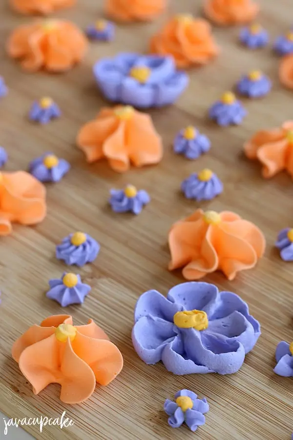 Close-up of completed Royal Icing flowers in purple and orange. 1 large purple, 1 large orange and 3 small purple flowers display the different techniques used to create different styles of petal.