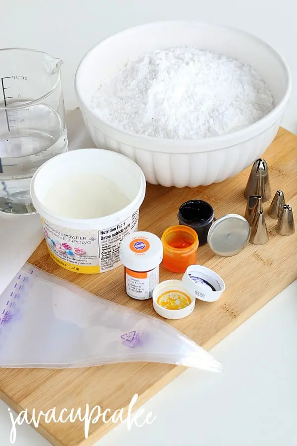 Ingredients for Royal Icing Flowers sitting on a wooden table. Large glass jug filled with water; white ceramic bowl filled with icing sugar; tub of meringue powder; food coloring in yellow, orange and purple; Wilton piping bags; Wilton piping tips in 5 different shapes.