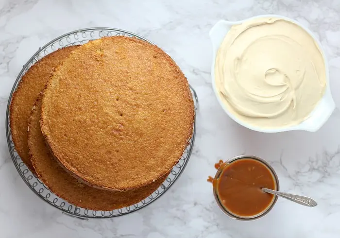 Some of the ingredients you need to make a Salted Caramel Cake. Caramel sponge cakes are stacked on top of eash other on a decorative glass cake stand. There is a small bowl of caramel  sauce with a teaspoon placed inside, next to that sits a large white bowl of caramel frosting.