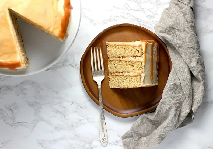 A slice of layer cake made from a caramel cake recipe is cut out of the rest of the cake. The slice lies side on a wooden plate with a fork. You can see the caramel drip down the side, and the 3 distinct layers of the cake. The rest of the cake is veiwed from above and you can see the salted caramel topping on the rest of the cake.