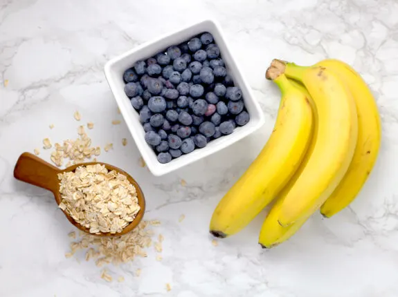 The ingredients that will be needed. Wooden spoon with rough cut oats, square whit bowl full of fresh whole blueberries, 3 ripe bananas.