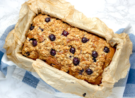 Freshly baked blueberry banana bread, the loaf has been removed from the oven but it remains in the tin with the parchment paper overlapping the edges.