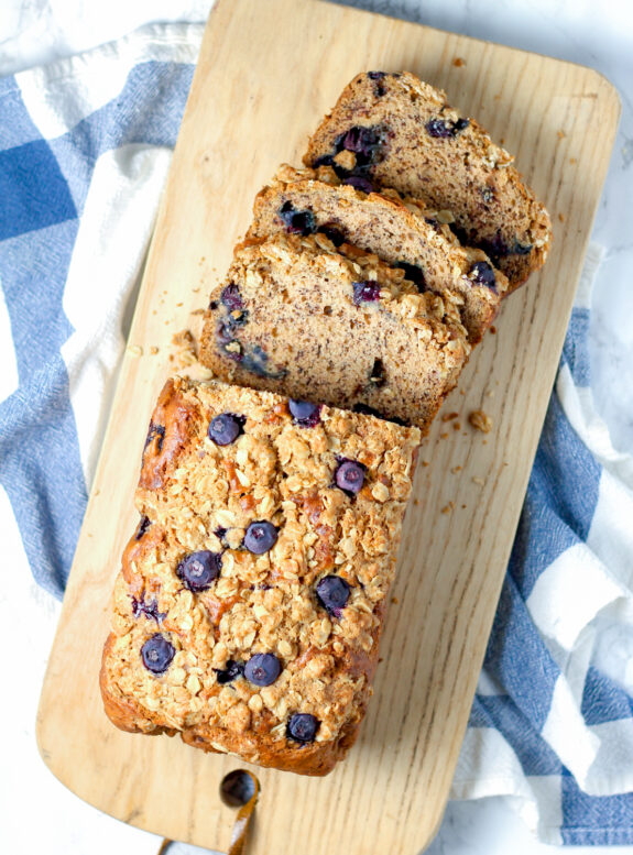 Blueberry Banana Bread with 3 slices cut from top. The loaf and slices are sitting on a wooden chopping board.