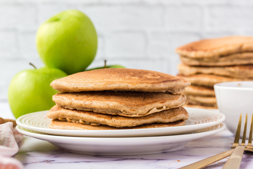 A small stack of Apple Pie Pancakes are siting on a white plate, and a large stack is in the background. There are 3 green apples next to the plate, alongside a gold fork.