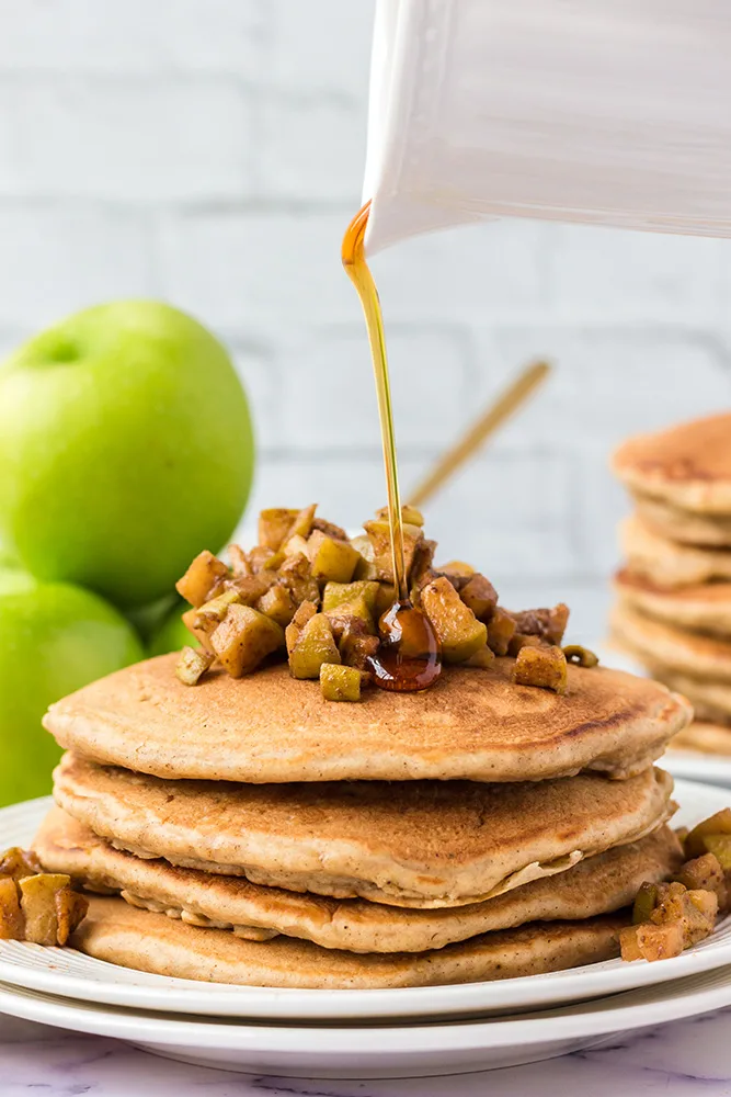 In a white kitchen, there is a stack of apple pie pancakes sitting on a white plate. the pancakes are topped with chopped, cooked apples and maple syrup is being drizzled on top. There is another stack of pancakes in the background alongside some green apples.