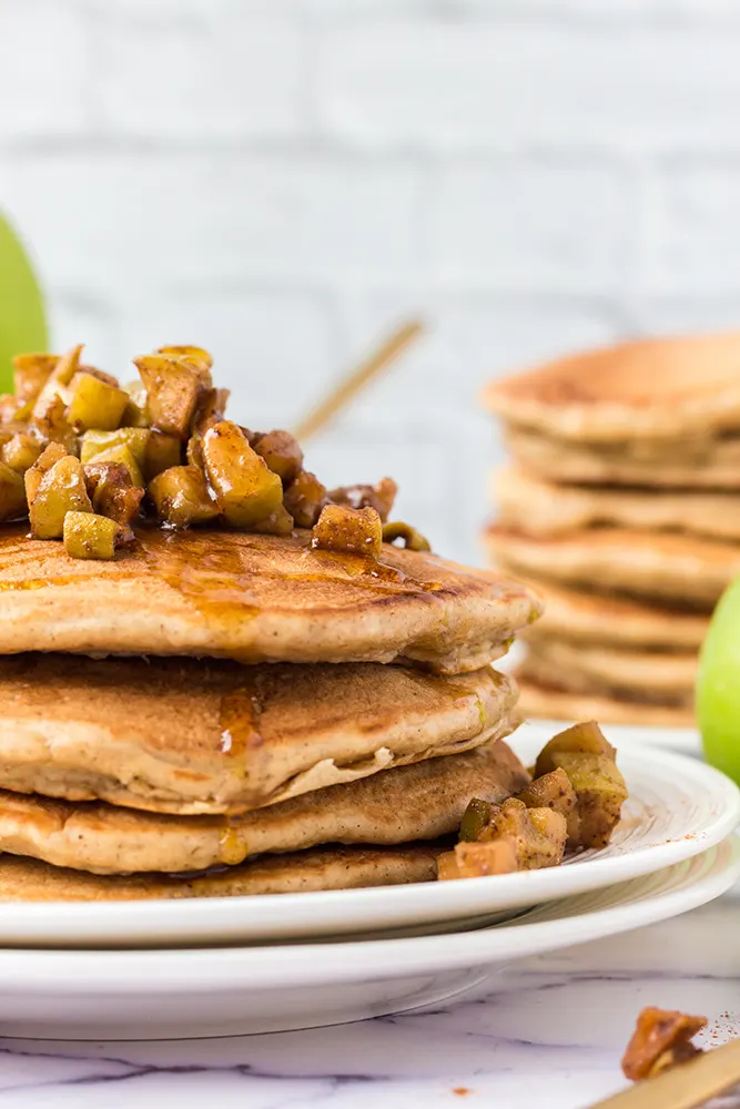 In a white kitchen there is a stack of apple cinnamon pancakes topped with stewed apples and maple syrup. The edge of the plate is outside of the frame so only half of the stack can be seen. In the background there is another large stack of apple pancakes.