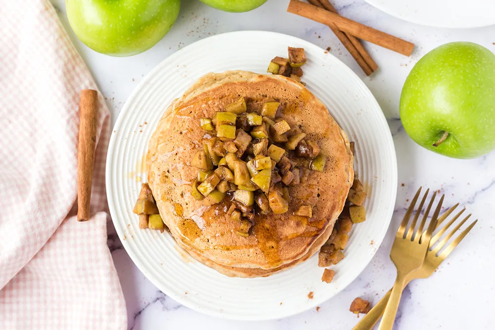View from above of easy apple pancakes. There is a stack on a white plate and they have been topped with cooked apples and maple syrup.
On top of the white marble counter is some gold forks, green apples, cinnamon sticks and a pink and white dish cloth.