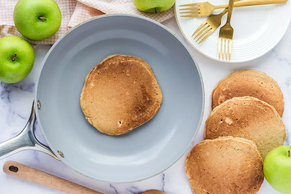 In a grey colored saucepan sits 1 apple pancake. Next to the pan is 3 more cooked pancakes. There is also a white plate with 3 gold forks on top. Some apples are left on top of the counter surrounding the other items.