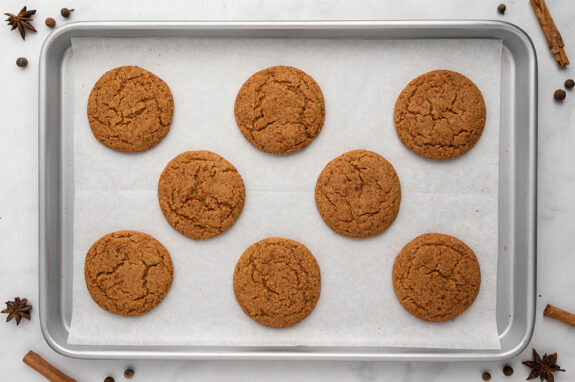 A baking tray lined with greaseproof paper has 8 cooked pumpkin spice cookies on top. There are a few spices like cinnamon stick and star anise laying around the counter.