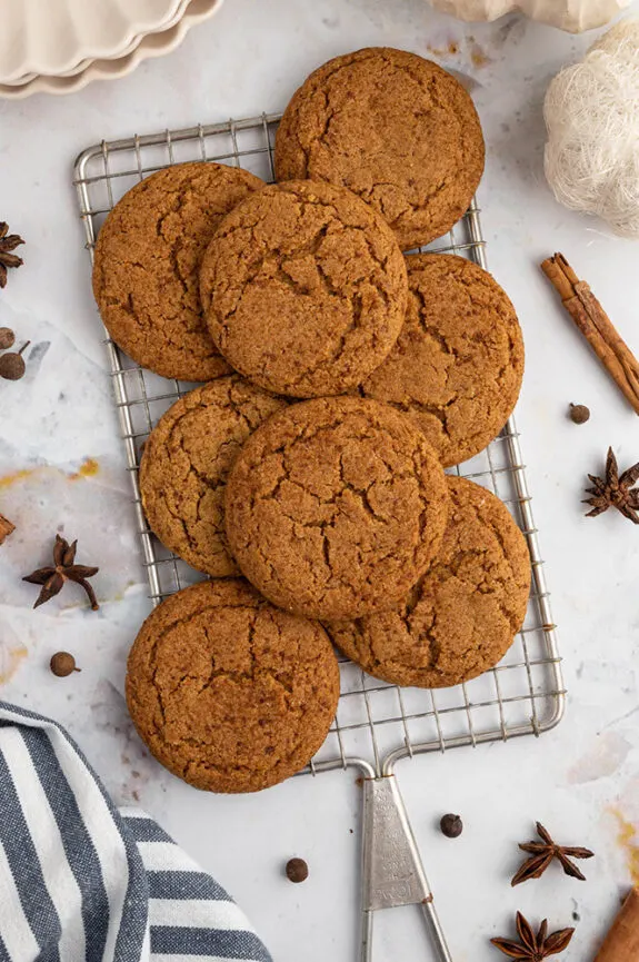 A white marble counter with a rectangle wire rack with lots of Fall pumpkin cookies on top. The spices used inside of the recipe like cinnamon and cloves are laying around the cookies on the counter.