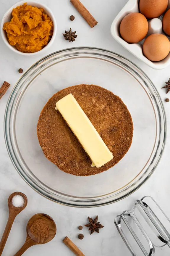 Some of the ingredients required to make these gluten free fall cookies. On a white counter top, there is a glass bowl filled with coconut sugar, a stick of soft, but not melted butter is sitting on top. There is a small white bowl filled with pumpkin puree and a carton of eggs can be seen in the corner, but you cannot see the full carton. 
Some wooden spoons are resting with some spice in them, like pumpkin spice and salt and more spices are scattered across the work space. In the bottom corner you can see the metal beaters for the electric whisk