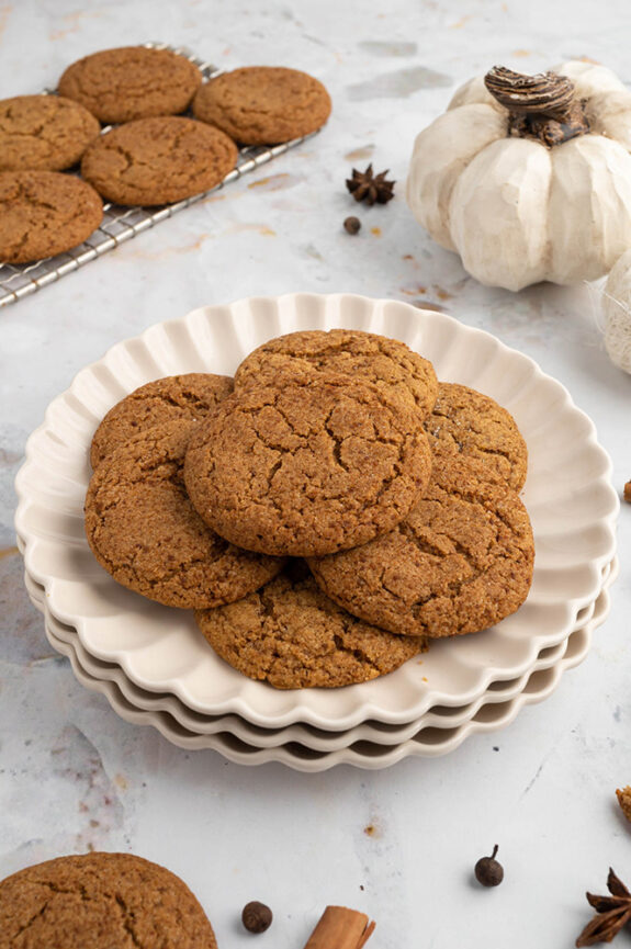 A white plate sitting on a white marble counter has 7 pumpkin cookies on top, spread out like a flower. In the background there is a wire rack with the extra cookies cooling on top.