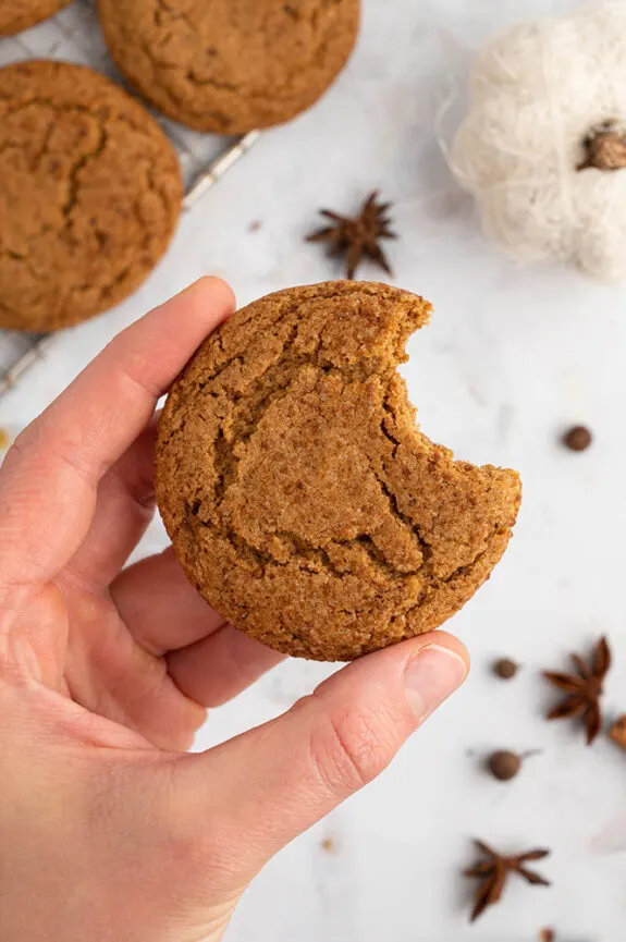 A hand holds a chewy gluten free pumpkin cookie that has had a bite taken out of it. In the background, on the counter top, the cooling rack is sitting with extra cookies on top of it. Some spices are scattered on the counter too.
