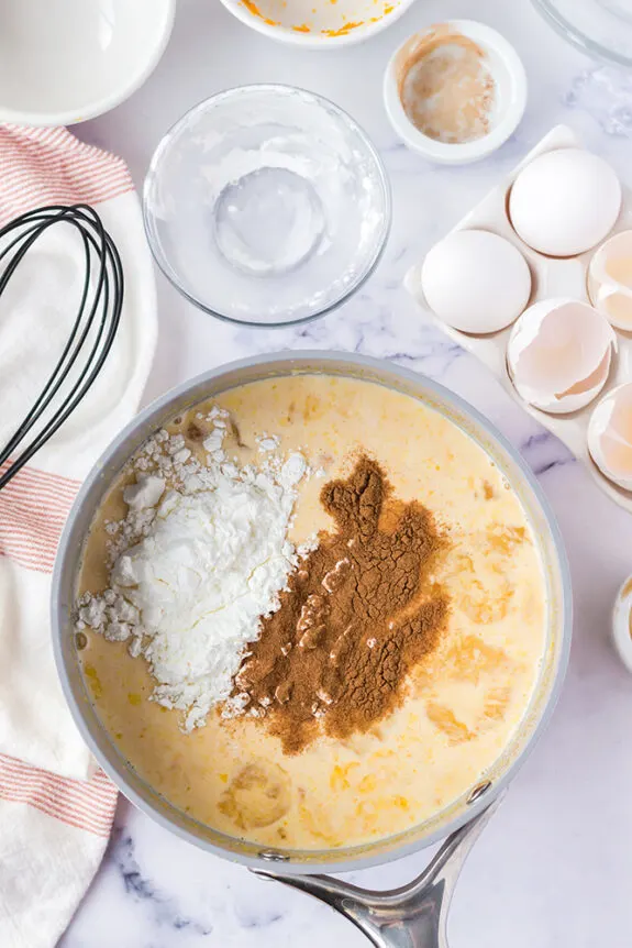 In a white kitchen, the dirty empty ramekin dishes that contained ingredients are now empty. There is a silver saucepan filled with pudding, spices have been added to the top of it but have not yet been incorporated. 