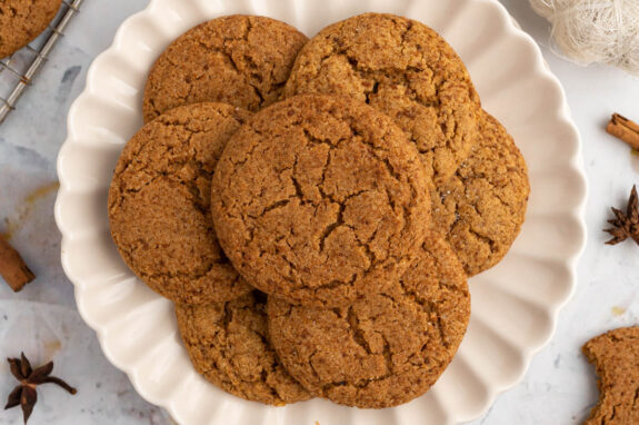 On a white plate, on a white counter, 7 gluten free pumpkin cookies sitting on a white plate, spread out like a flower. In the background there are some leftover spices and cookie crumbs.