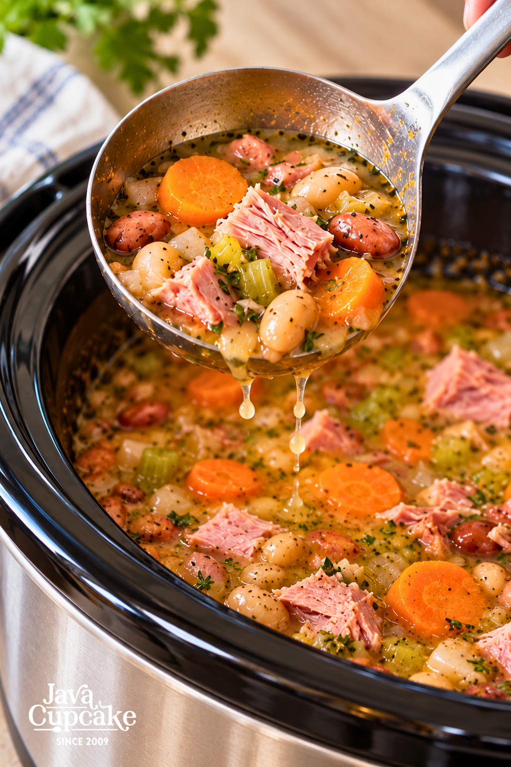 Close-up of a ladle filled with ham, mixed beans, carrots, and celery above a slow cooker of soup.
