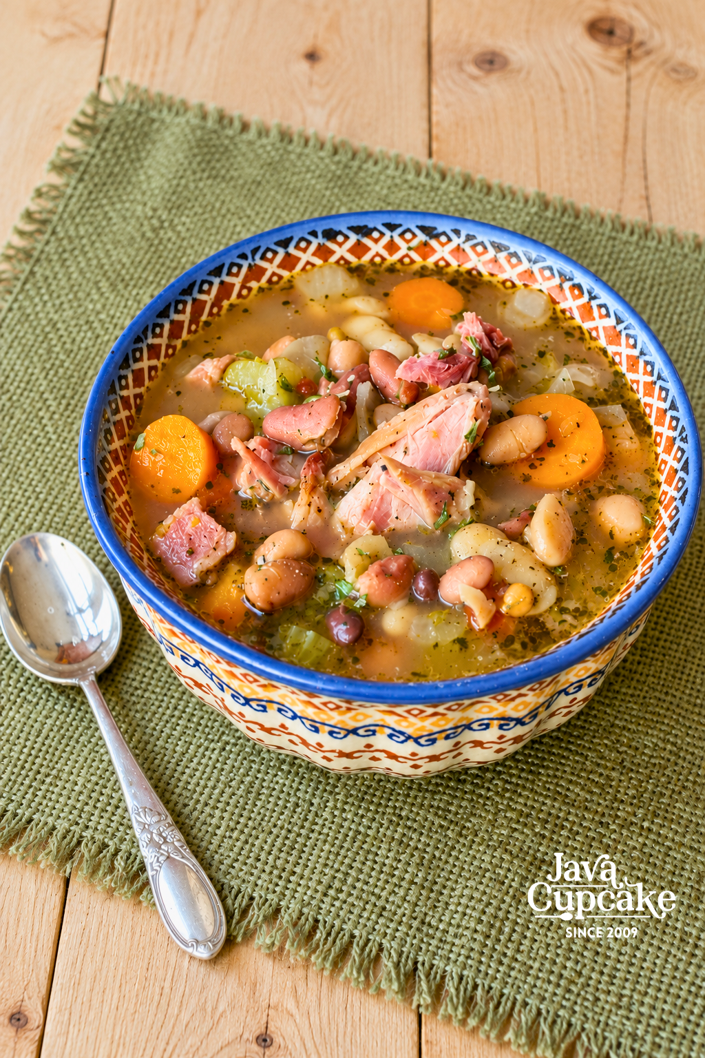 Ham hock and 15 bean soup served in a Polish pottery bowl with a silver spoon on a green woven cloth.